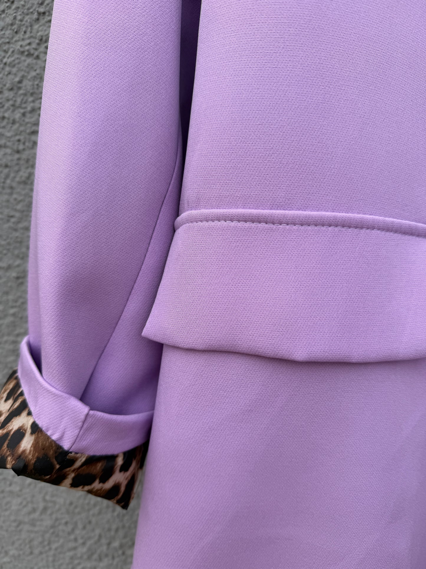 Eine Nahaufnahme des Eleganter Nice Fit Blazers in flieder mit Leopardenprint von Feinraummode zeigt die Klappe und den umgeschlagenen Ärmel, der das Futter mit Leopardenprint vor einem strukturierten grauen Hintergrund enthüllt.