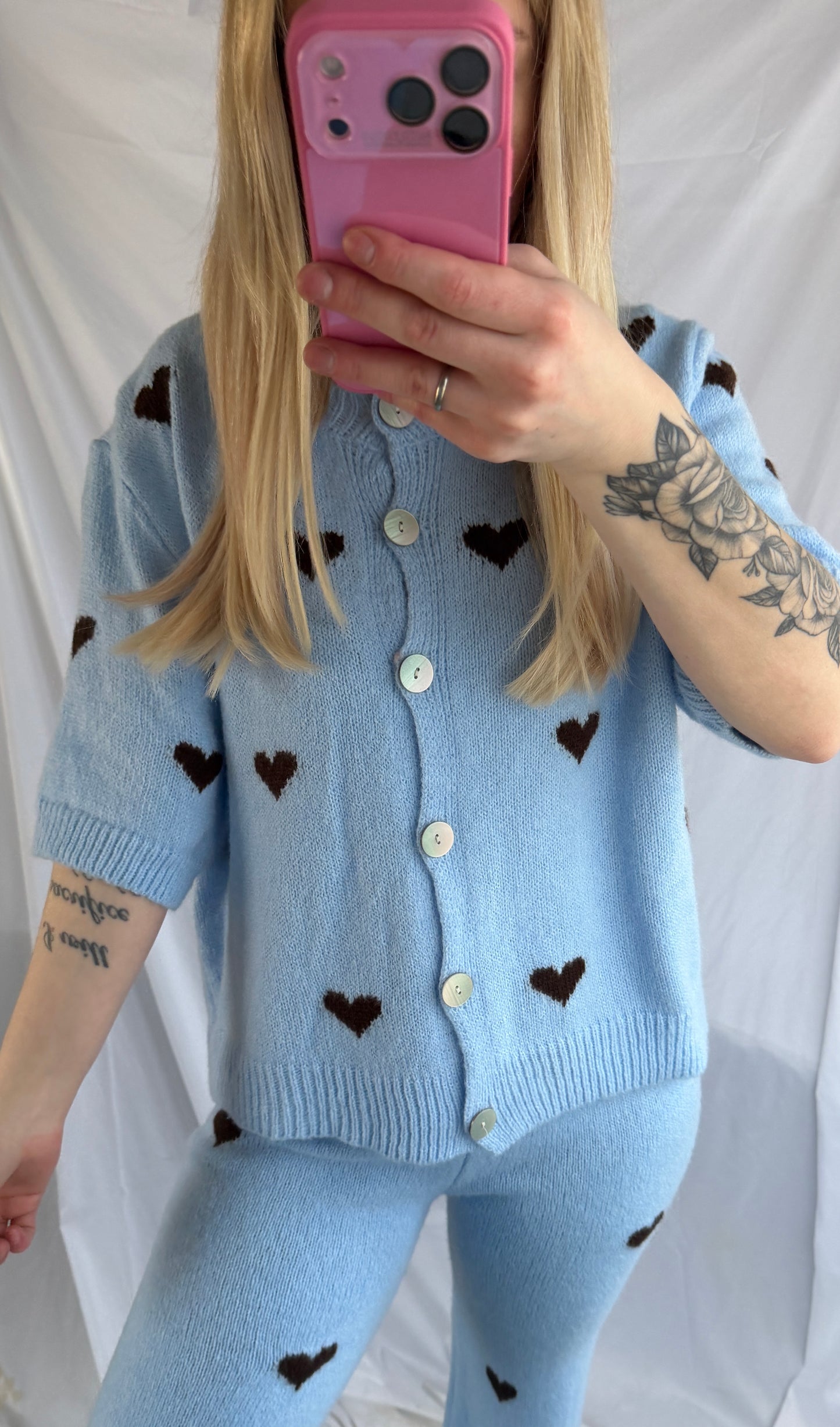 Eine Person mit langen blonden Haaren macht ein Selfie im Spiegel und trägt das Feinraummode Cozy Set Hearts (Lounge Anzug Herzen), bestehend aus einer bequemen Hose mit dunklem Herz-Print und einer hellblauen Strickjacke mit kurzen Ärmeln. Auf ihrem linken Arm sind Blumen- und Zitat-Tattoos zu sehen. Rosa Handytasche.