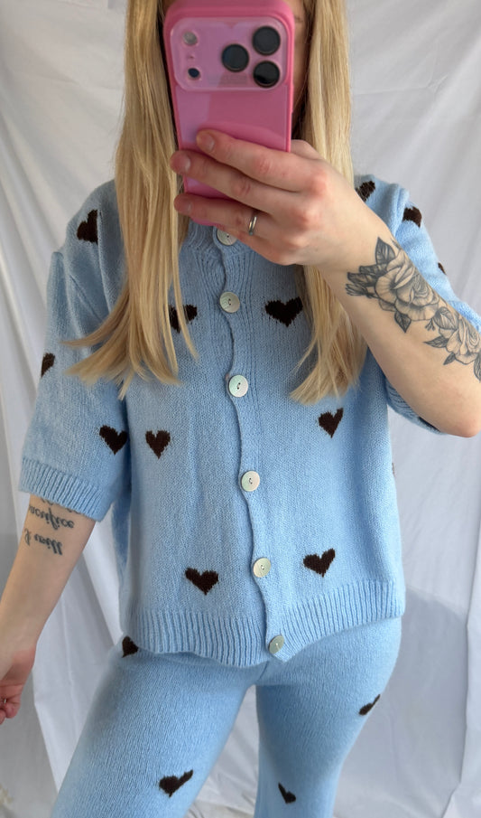 Eine Person mit langen blonden Haaren macht ein Selfie im Spiegel und trägt das Feinraummode Cozy Set Hearts (Lounge Anzug Herzen), bestehend aus einer bequemen Hose mit dunklem Herz-Print und einer hellblauen Strickjacke mit kurzen Ärmeln. Auf ihrem linken Arm sind Blumen- und Zitat-Tattoos zu sehen. Rosa Handytasche.