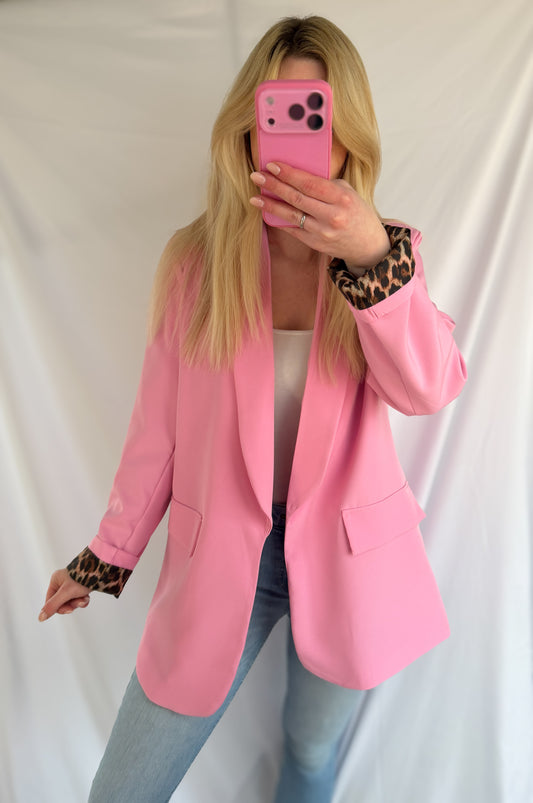 Eine Person mit langen blonden Haaren trägt den Eleganter Nice Fit Blazer in rosa mit Leopardenprint von Feinraummode, gepaart mit einem weißen Top und hellblauen Jeans, und macht ein Spiegel-Selfie mit einem rosafarbenen Telefon vor einem weißen Hintergrund.