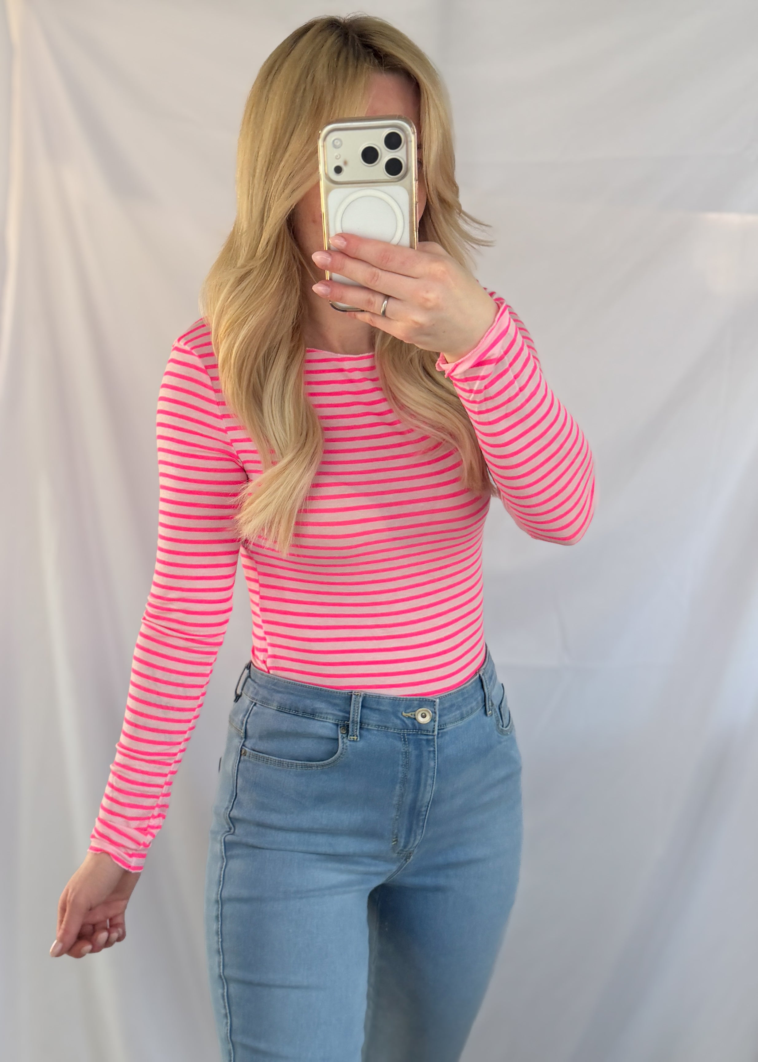 Eine Person mit langen blonden Haaren trägt das Feinraummode Gestreiftes Longsleeve mit Viskose & Cashmere in Rosa, gepaart mit hochgezogenen Bluejeans, und macht ein Spiegelselfie mit einem Smartphone. Weißer Stoff als Hintergrund.