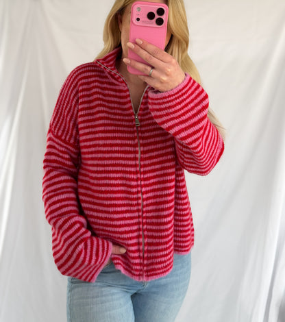 Eine Person mit langen blonden Haaren trägt die Feinraummode Gestreifter Zip Cardigan rot und hellblaue Jeans, hält sich ein rosa Telefon vor das Gesicht und steht vor einem schlichten weißen Hintergrund.