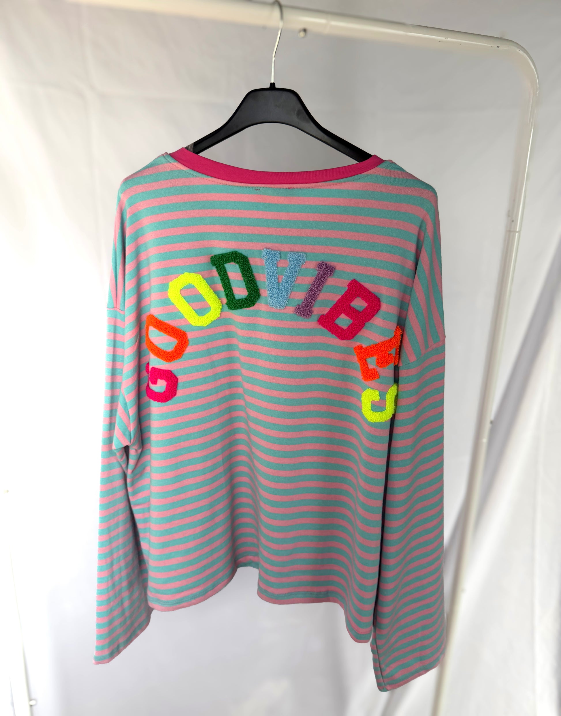 Das B-Ware - Good Vibes Shirt gestreift von Feinraummode ist ein übergroßes Longsleeve mit hellblauen und rosa Streifen, das auf einem schwarzen Bügel hängt. "GOOD VIBES" Stickerei in bunten, fetten Buchstaben ziert den Rücken.