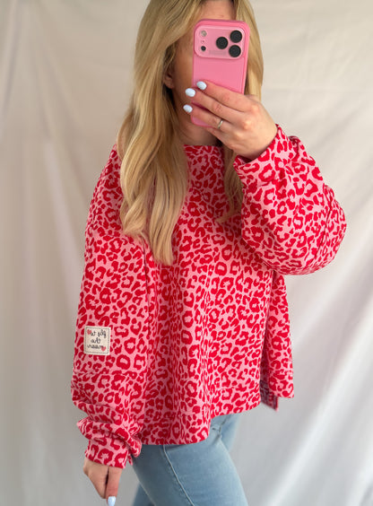 Eine Person mit langen blonden Haaren trägt das Feinraummode Oversize Sweatshirt im Leoparden Look - rot und blaue Jeans und hält ein pinkes Handy für ein Spiegel-Selfie vor einem schlichten hellen Laken.