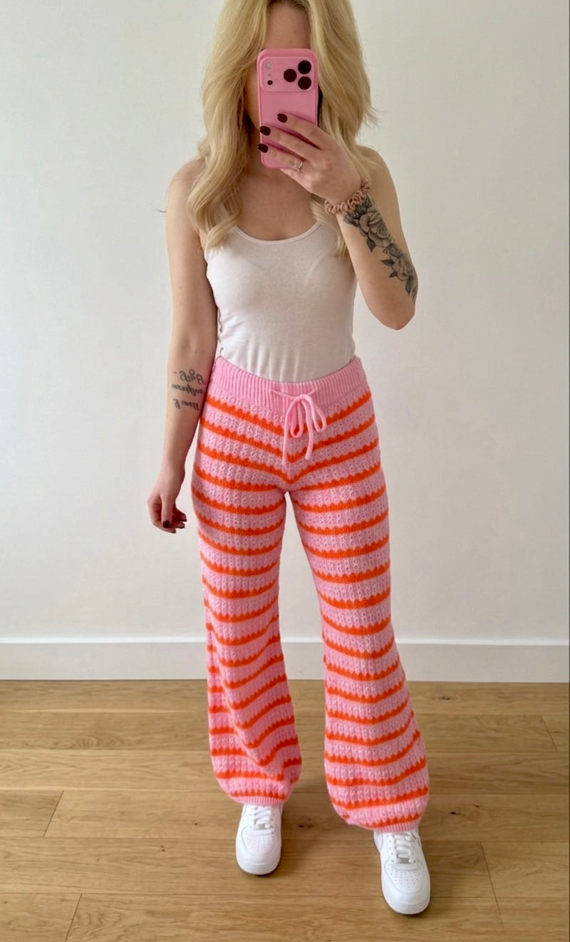 Eine Person mit langen blonden Haaren macht ein Spiegel-Selfie in einem weißen Tanktop und der Kuschelig gestreiften Strickhose im Wellen-Look - Pink & Orange von Feinraummode, gestylt mit weißen Turnschuhen auf einem Holzboden vor einer schlichten weißen Wand.