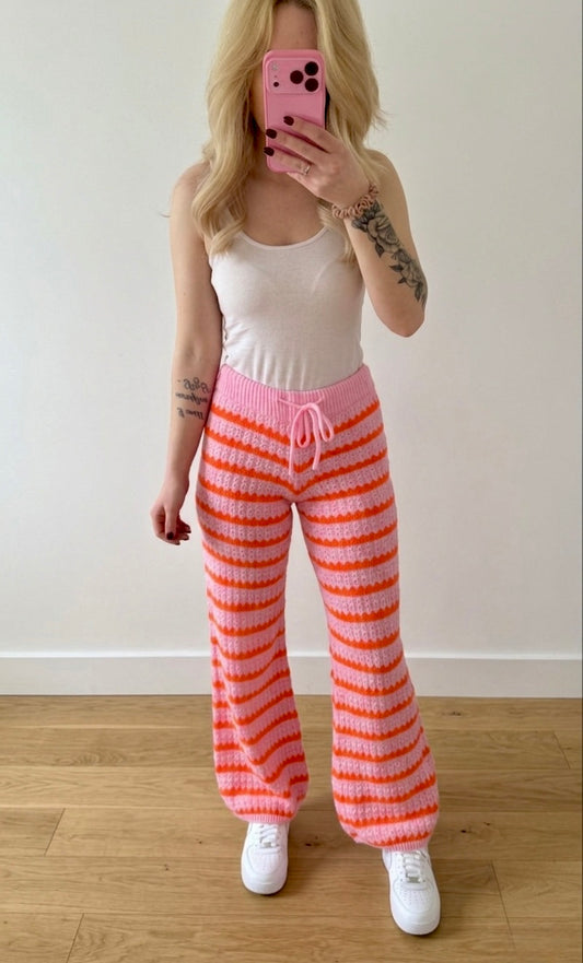 Eine Person mit langen blonden Haaren macht ein Spiegel-Selfie in einem weißen Tanktop und der Kuschelig gestreiften Strickhose im Wellen-Look - Pink & Orange von Feinraummode, gestylt mit weißen Turnschuhen auf einem Holzboden vor einer schlichten weißen Wand.