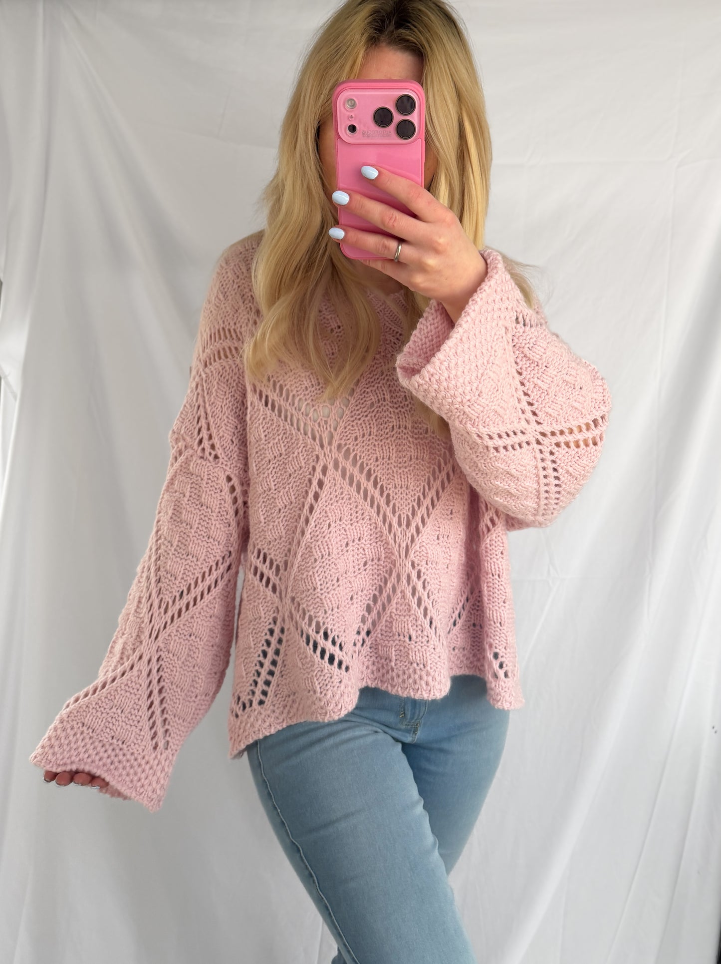 Eine Frau mit langen blonden Haaren trägt den Oversize Pullover Christina - Damen Strickpullover rosa im lässigen Oversize Look von Feinraummode und hellblaue Jeans und macht ein Spiegel-Selfie mit einem rosafarbenen Handy vor einem schlichten weißen Laken-Hintergrund.