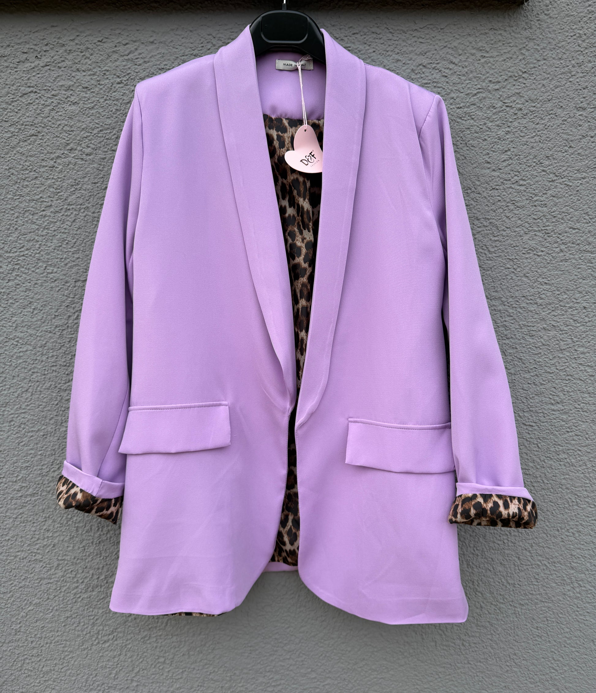 Der elegante Nice Fit Blazer in Flieder mit Leopardenprint von Feinraummode hängt an einem schwarzen Bügel vor grauer Wand, die Ärmel sind hochgekrempelt und am Kragen befindet sich ein herzförmiges Etikett.