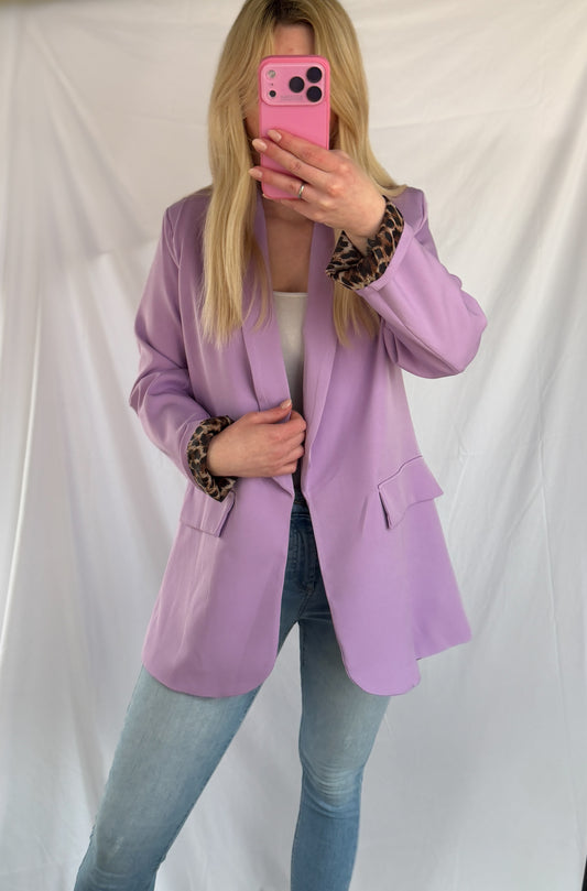 Eine Person mit langen blonden Haaren trägt den Feinraummode Eleganter Nice Fit Blazer in flieder mit Leopardenprint, gepaart mit hellblauen Jeans und einem weißen Oberteil, und macht ein Spiegel-Selfie mit einem rosafarbenen Telefon vor einem schlichten weißen Hintergrund.