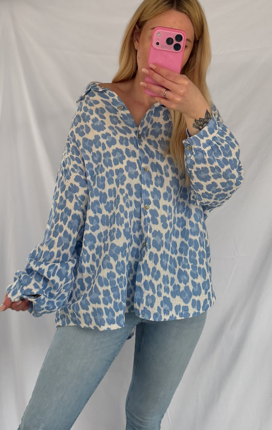 Eine Person mit langen blonden Haaren macht ein Spiegel-Selfie vor einem schlichten weißen Hintergrund. Sie trägt die Oversize Musselinbluse Leopardenmuster blau weiß von Feinraummode, gepaart mit hellblauen Jeans.