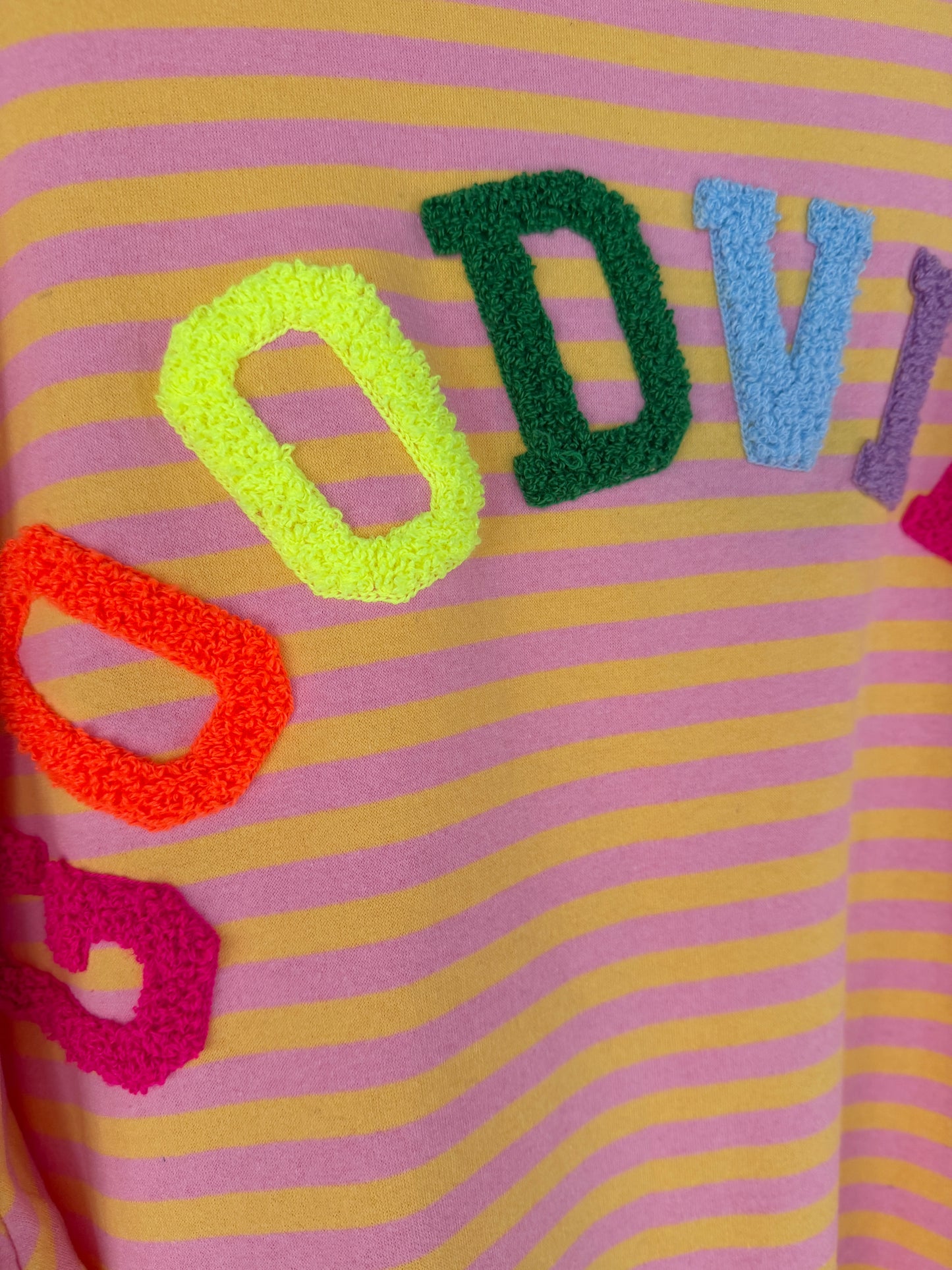 Eine Nahaufnahme des Good Vibes Streifen Longsleeve von Feinraummode zeigt die pinkfarbenen und gelben Streifen mit bunten, strukturierten Buchstaben - rot, gelb, grün und blau - die teilweise ein Wort über dieses trendige Stück für Frühlingsmode buchstabieren.