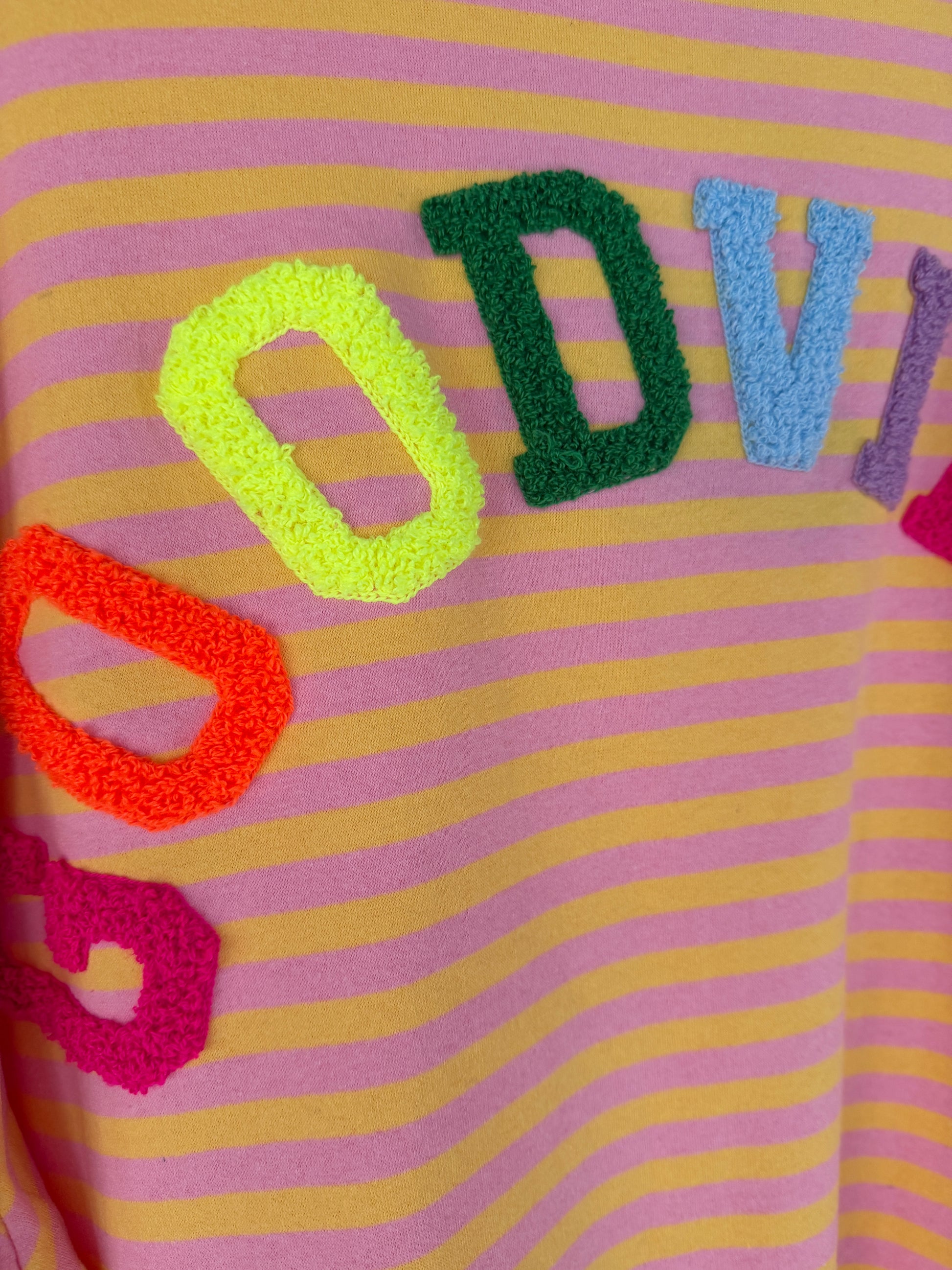 Eine Nahaufnahme des Good Vibes Streifen Longsleeve von Feinraummode zeigt die pinkfarbenen und gelben Streifen mit bunten, strukturierten Buchstaben - rot, gelb, grün und blau - die teilweise ein Wort über dieses trendige Stück für Frühlingsmode buchstabieren.