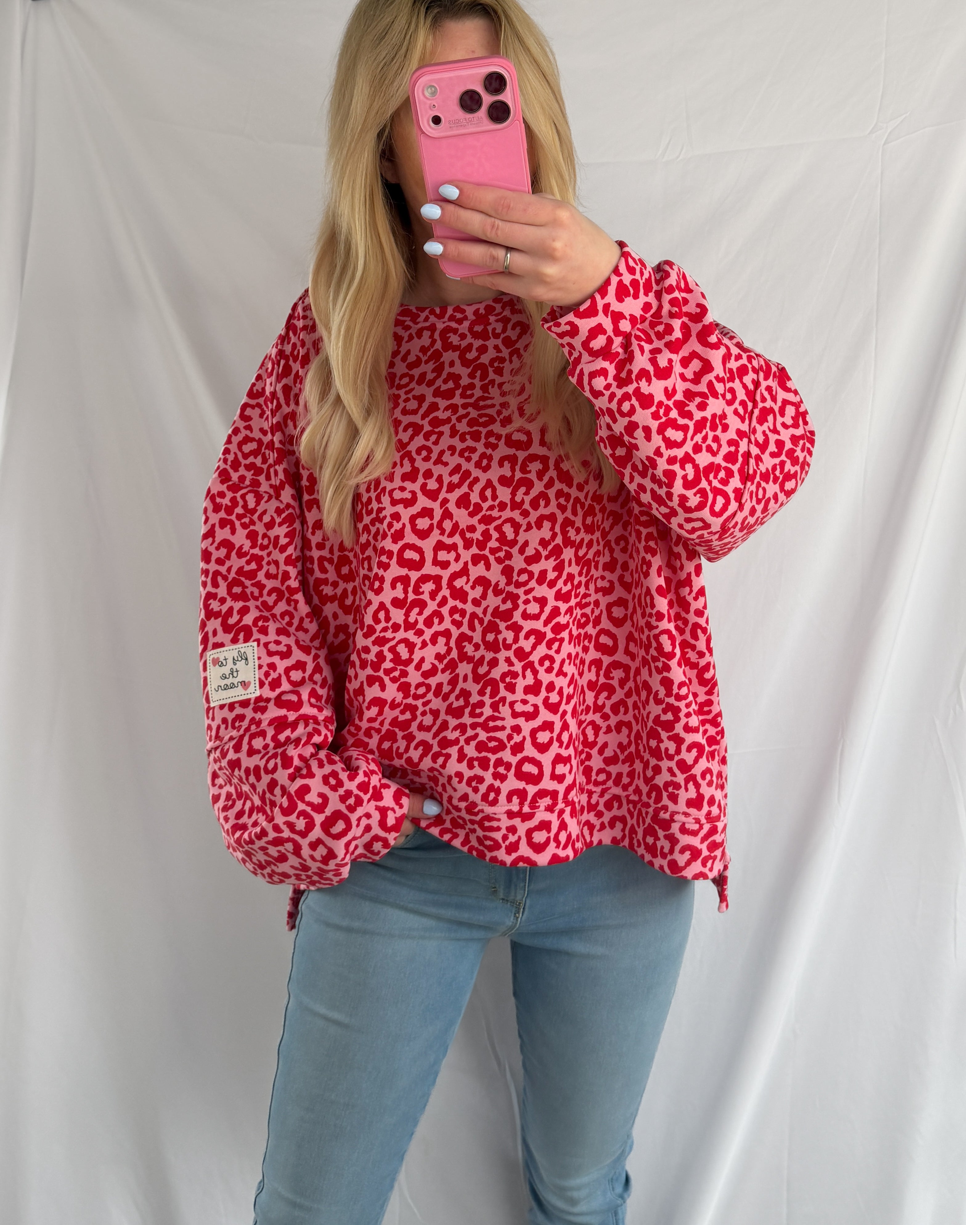 Eine Person mit langen blonden Haaren macht ein Selfie im Spiegel. Sie trägt das Feinraummode Oversize Sweatshirt im Leoparden Look - rot und hellblaue Jeans und steht vor einem schlichten weißen Hintergrund.