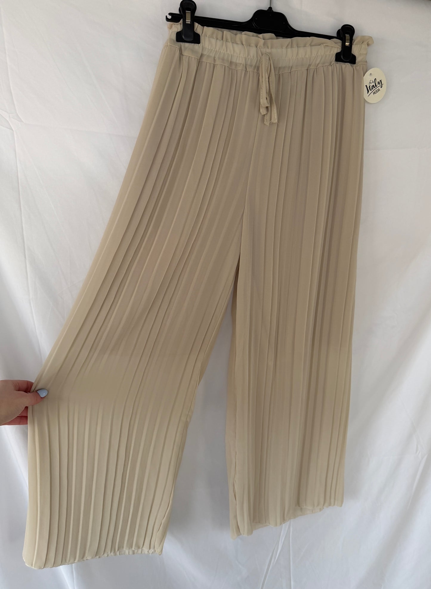 Eine hellbeige Plissee-Hose Amanda von Feinraummode hängt auf einem schwarzen Bügel vor einem weißen Hintergrund. Eine Hand hält ein weites Bein und hebt die fließenden Falten und das Preisschild am Gummibund hervor.