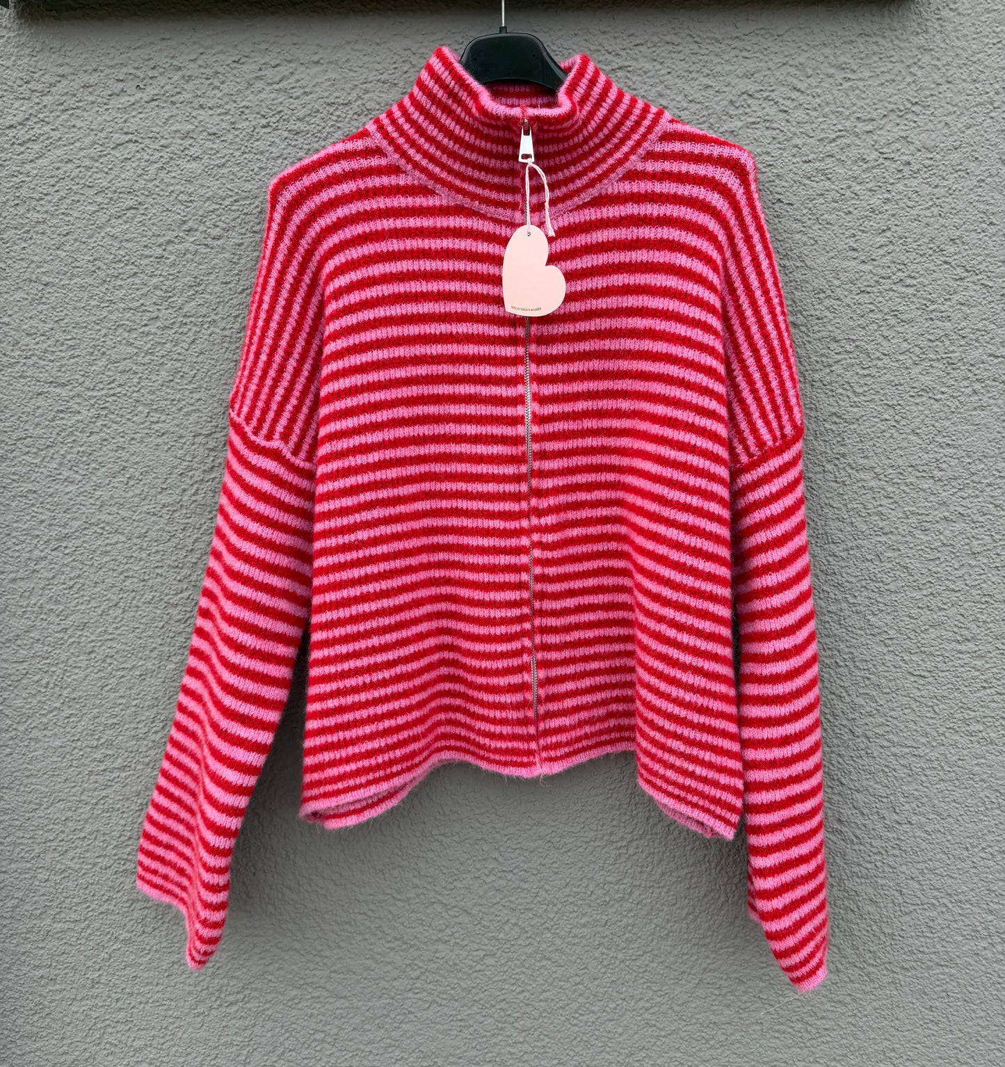 Der Gestreifter Zip Cardigan rot von Feinraummode mit pinken Streifen und hohem Kragen hängt an einem schwarzen Bügel vor einer grau strukturierten Wand.