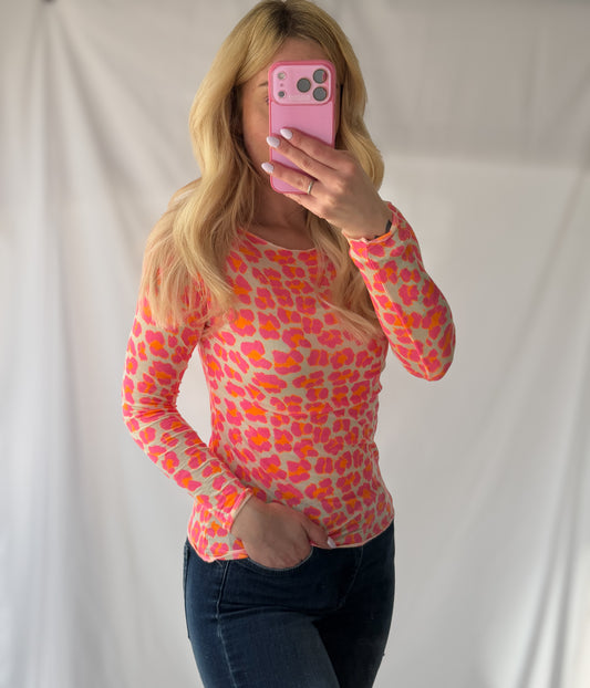 Eine Person mit langen blonden Haaren macht ein Selfie im Spiegel und trägt dabei die Feinraummode Longsleeve Leoparden Muster orange mit dunkler Jeans. Der Hintergrund ist einfarbig und hell gefärbt.