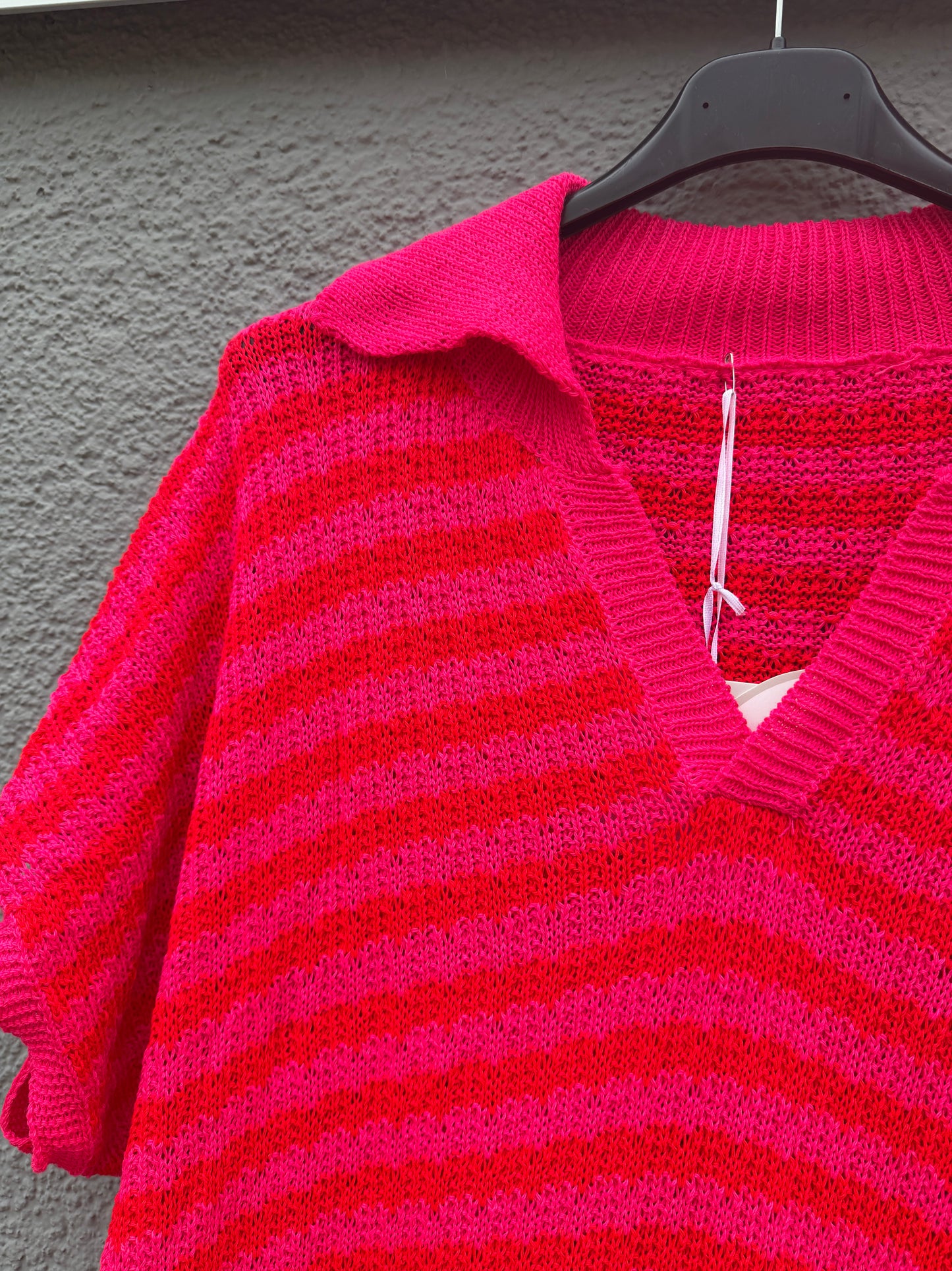 Eine Nahaufnahme des Sommerlichen Strickkleids mit Streifen pink von Feinraummode, das auf einem schwarzen Bügel vor einer grau strukturierten Wand hängt.