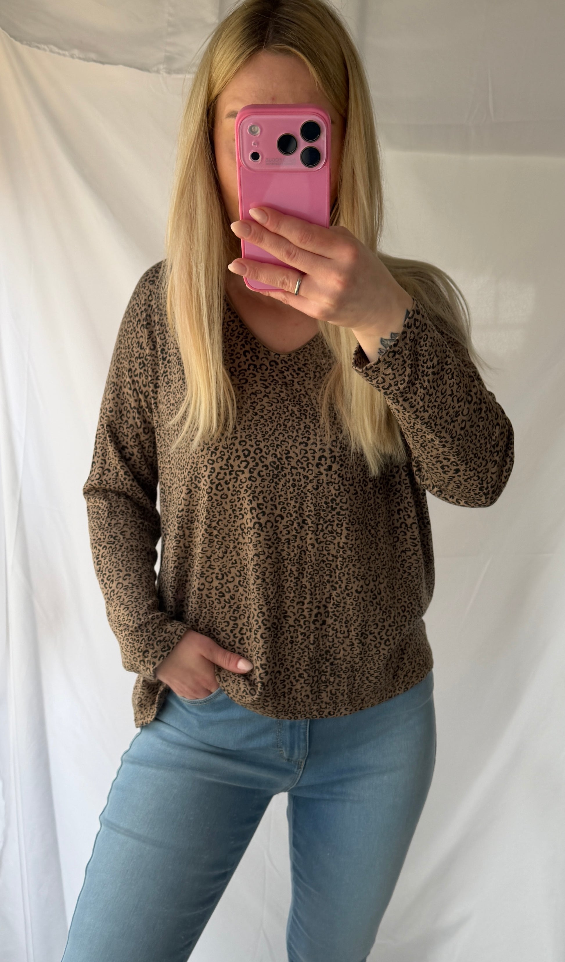 Eine Frau mit langen blonden Haaren macht ein Spiegel-Selfie in einem Feinraummode Leo Pullover Damen Animalprint - 20% Angora Wolle, gepaart mit hellblauen Jeans. Sie hält ein rosafarbenes Telefon, das ihr Gesicht teilweise verdeckt; dahinter befindet sich ein weißer Hintergrund.