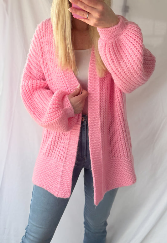 Eine Person mit langen blonden Haaren modelliert den Kuscheliger Cardigan Lara von Feinraummode in pinkem Grobstrick über einem weißen Top und Jeans und macht ein Selfie vor einem schlichten weißen Hintergrund. Perfekt für Frühling & Herbst, Einheitsgröße.