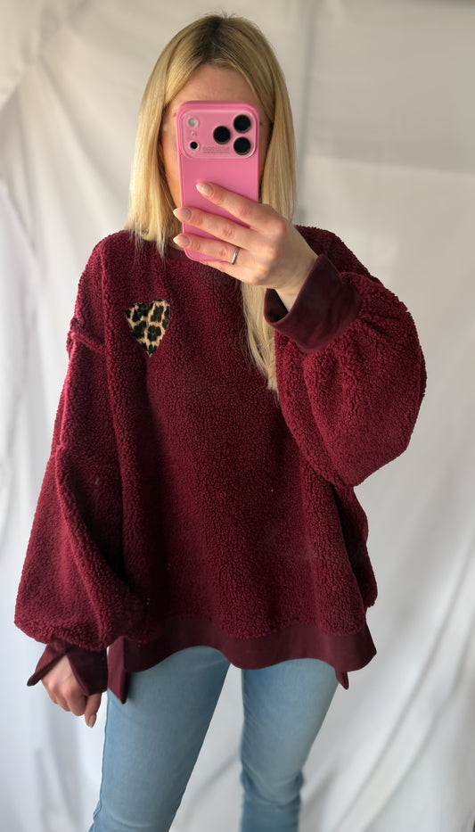 Eine Person mit langen blonden Haaren trägt den Flauschigen Teddypullover Julia von Feinraummode mit einem Herzaufnäher im Leo-Look, kombiniert mit einer hellblauen Jeans, und hält ein rosafarbenes Handy in der Hand, das ihr Gesicht teilweise verdeckt.