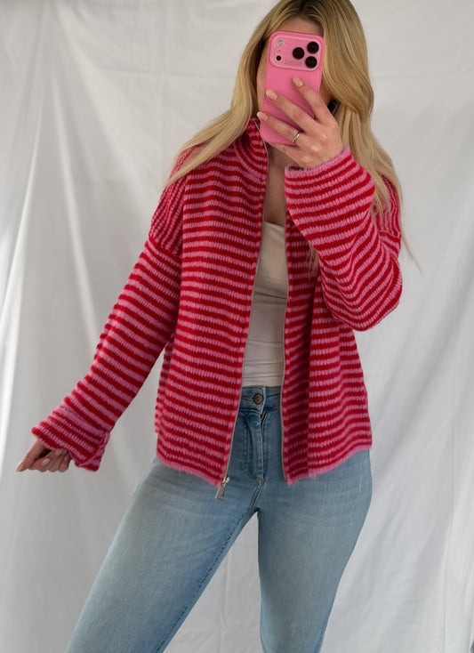 Eine Person mit langen blonden Haaren trägt die Feinraummode Gestreifter Zip Cardigan rot über einem weißen Oberteil und hellblauen Jeans und hält sich vor einem weißen Hintergrund ein rosa Telefon vors Gesicht.