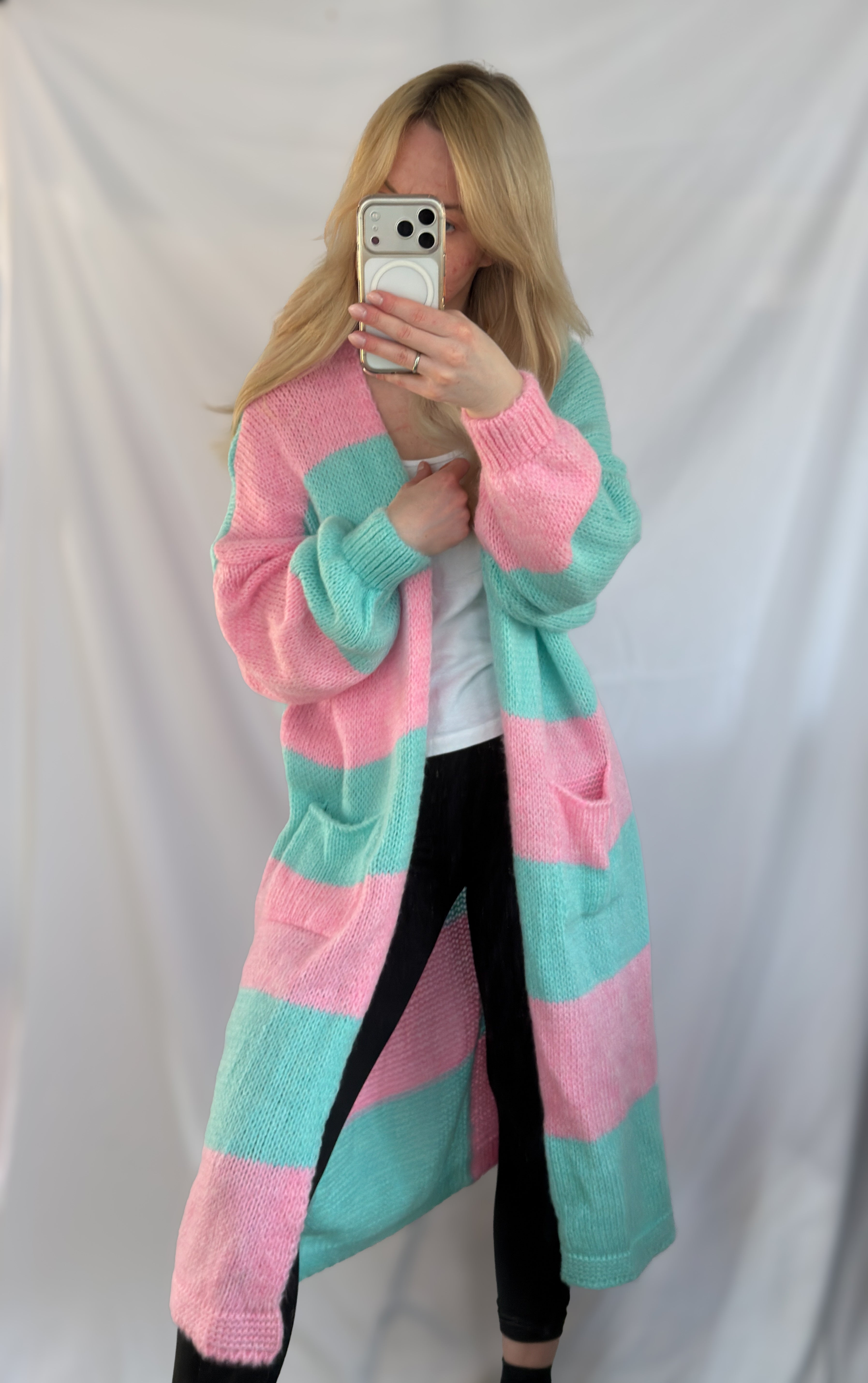 Eine Person mit langen blonden Haaren macht ein Selfie im Spiegel und trägt dabei den Feinraummode Langer Oversize Cardigan mit Blockstreifen - rosa blau in Einheitsgröße über einem weißen Oberteil und einer schwarzen Hose, vor einem weißen Hintergrund. Der Look ist gemütlich und stilvoll.