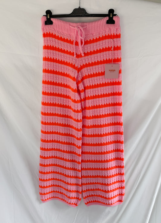 Die Kuschelig gestreifte Strickhose im Wellen-Look - Pink & Orange von Feinraummode mit weitem Bein, Kordelzug in der Taille und HANA-Tag wird auf einem schwarzen Bügel vor weißem Hintergrund präsentiert.