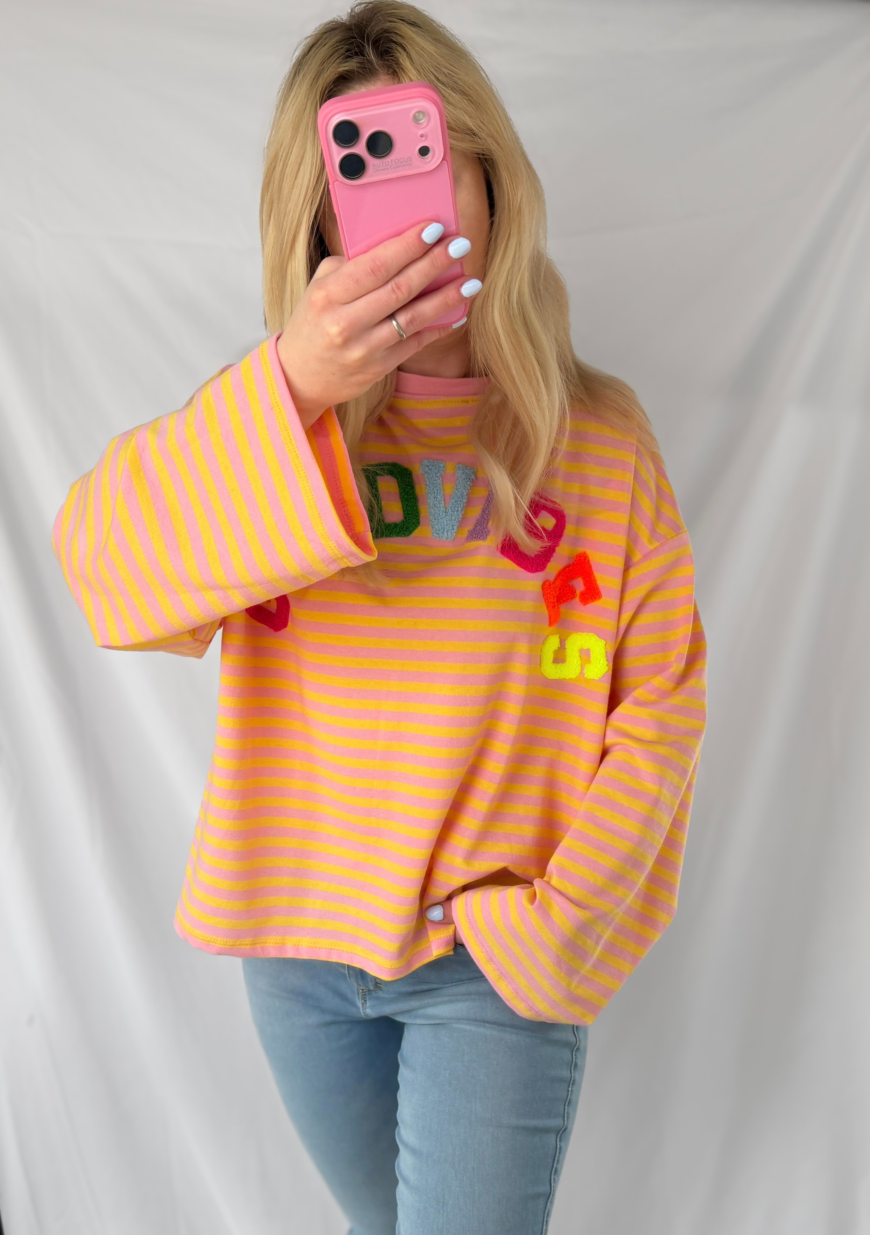 Eine Person mit langen blonden Haaren trägt das Feinraummode Good Vibes Streifen Longsleeve in rosa und gelben Streifen mit bunten Buchstaben, hellblaue Jeans und hält ein rosafarbenes Telefon - ein lässiger Frühlingslook vor weißem Hintergrund.