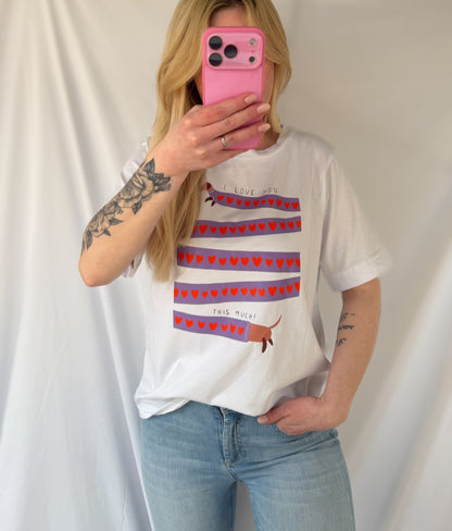 Eine Person mit langen blonden Haaren trägt die Feinraummode Weißes T-Shirt mit Dackelprint & Herzen und hellblaue Jeans und hält ein rosa Telefon vor einem weißen Stoffhintergrund, der die Passform des Shirts betont.