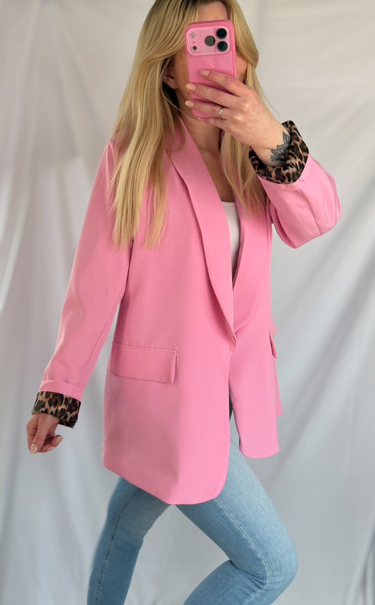 Eine Person mit langen blonden Haaren trägt den Feinraummode Eleganter Nice Fit Blazer in rosa mit Leopardenprint, gepaart mit hellblauen Jeans und einem weißen Oberteil, und macht ein Spiegel-Selfie mit einem rosafarbenen Telefon vor einem weißen Hintergrund.