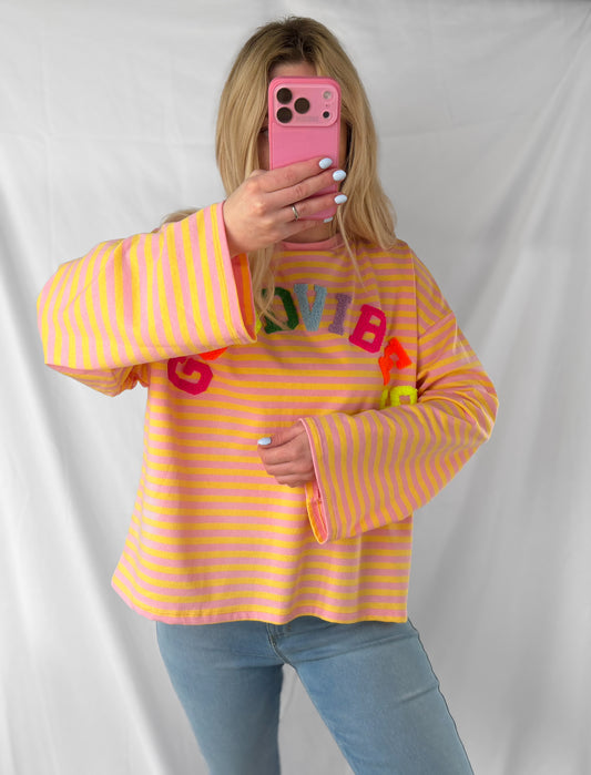 Eine Person mit langen blonden Haaren trägt das Feinraummode Good Vibes Streifen Longsleeve in Pink und Gelb mit fetten Schriftzügen und hellblauen Jeans und hält sich ein pinkes Handy vor das Gesicht. Ein schlichtes weißes Laken hinter ihr verbreitet perfekte Frühlingsmode-Vibes.