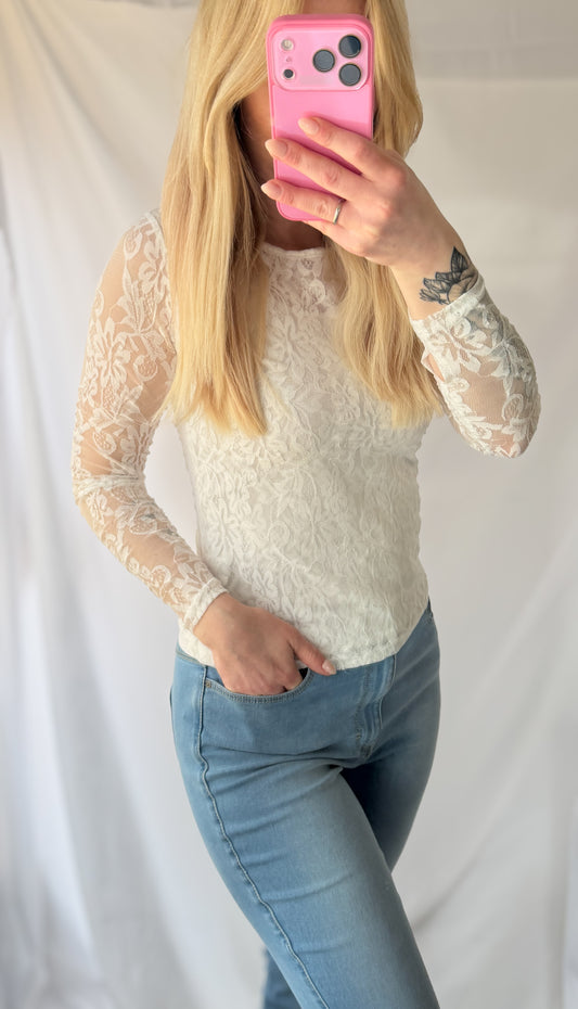 Eine Person mit langen blonden Haaren trägt die Feinraummode Shirt mit Spitze Pia in braun und weiß und hellblaue Jeans, steht vor einem weißen Hintergrund und hält sich ein rosafarbenes Handy vor das Gesicht.