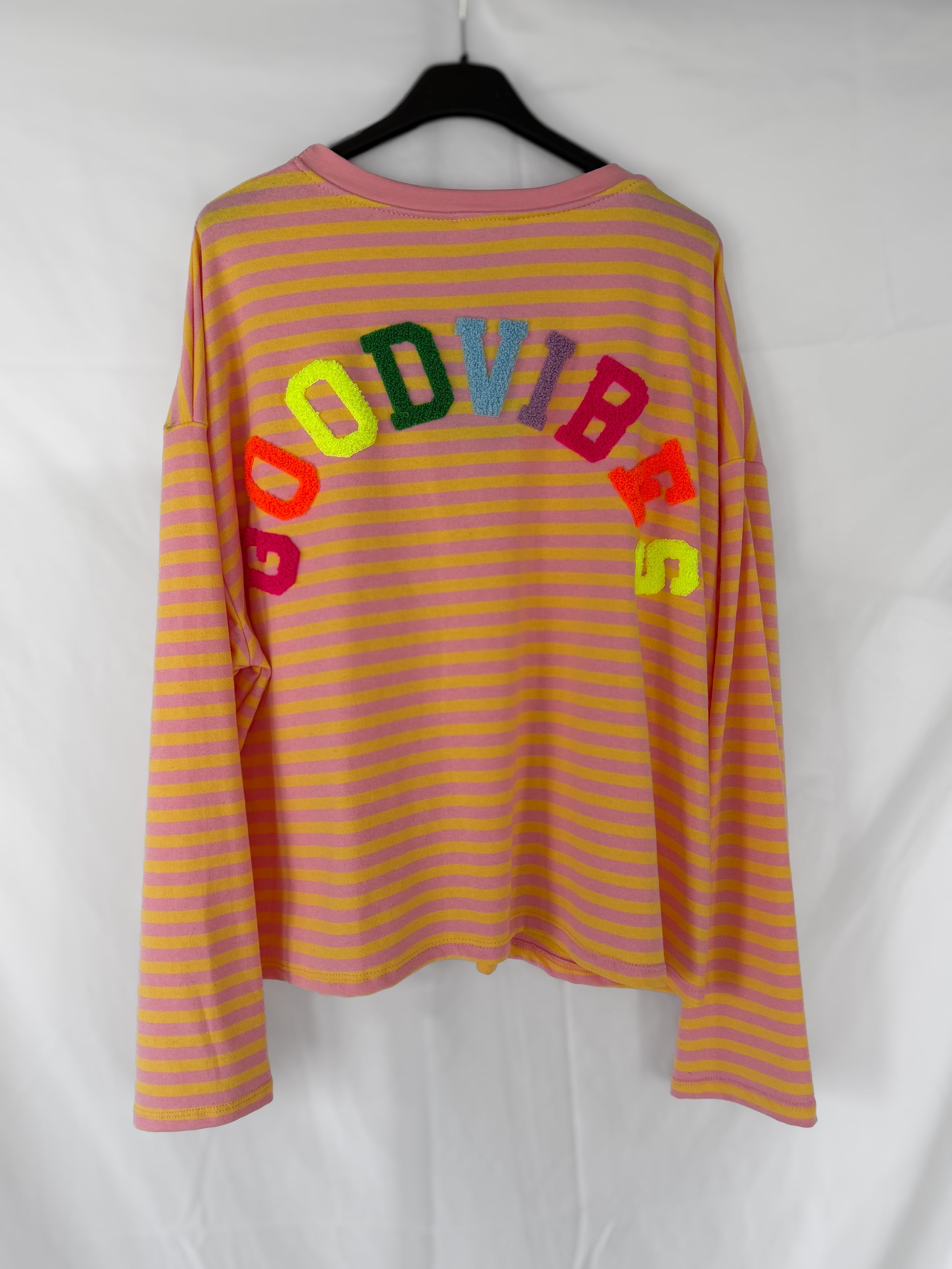 Das Feinraummode Good Vibes Streifen Longsleeve in rosa und gelben Streifen hängt an einem schwarzen Bügel. "GOOD VIBES" in bunten, strukturierten Buchstaben wölbt sich quer über den Rücken - ideal für Frühlingsmode.