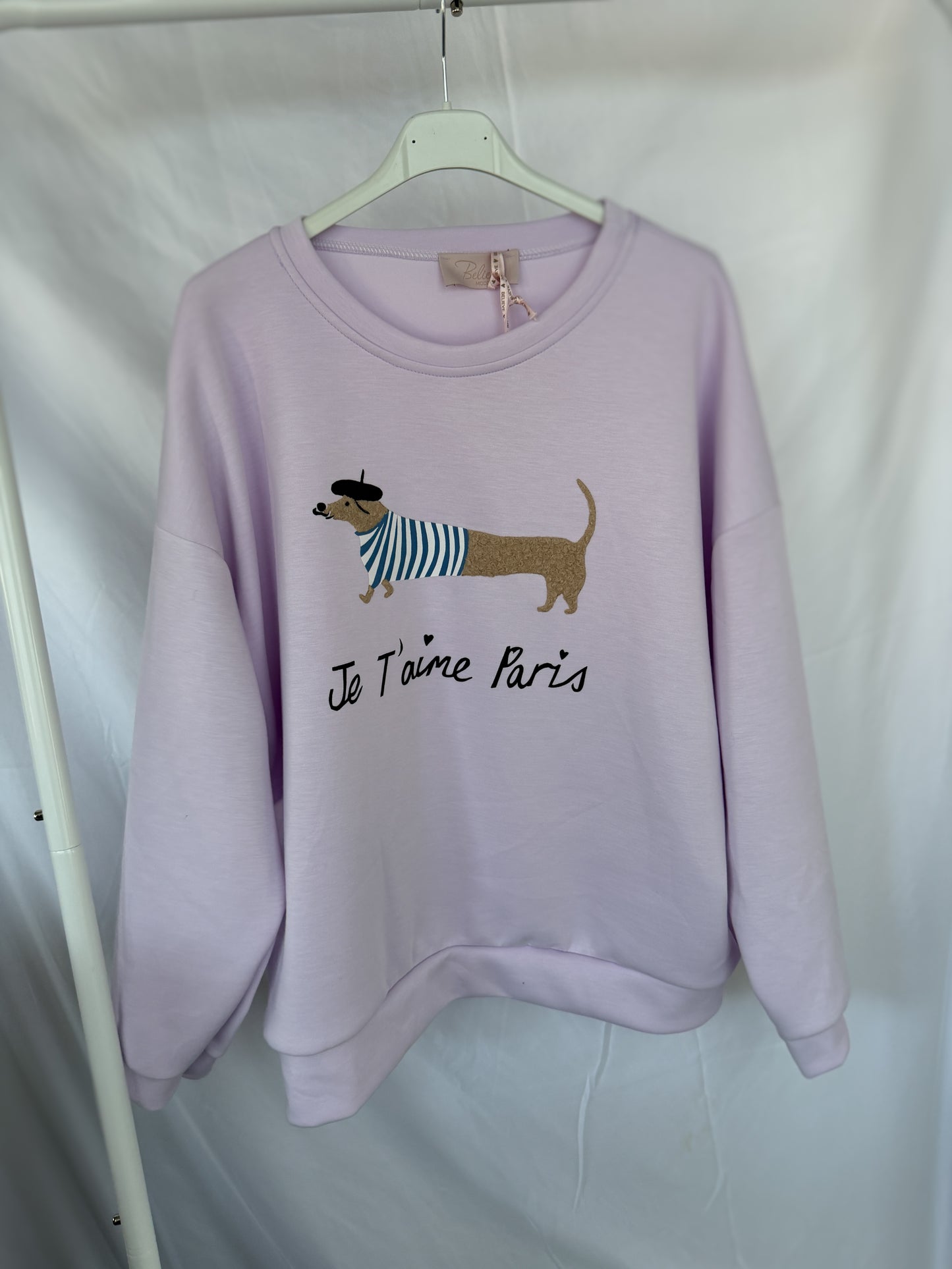 An einem weißen Kleiderbügel hängt ein hellvioletter Feinraummode Sweatshirt Dackelprint / Je t'aime Paris Schriftzug Flieder, der einen niedlichen Dackel mit Baskenmütze und gestreiftem Hemd zeigt, darunter "Je T'aime Paris".