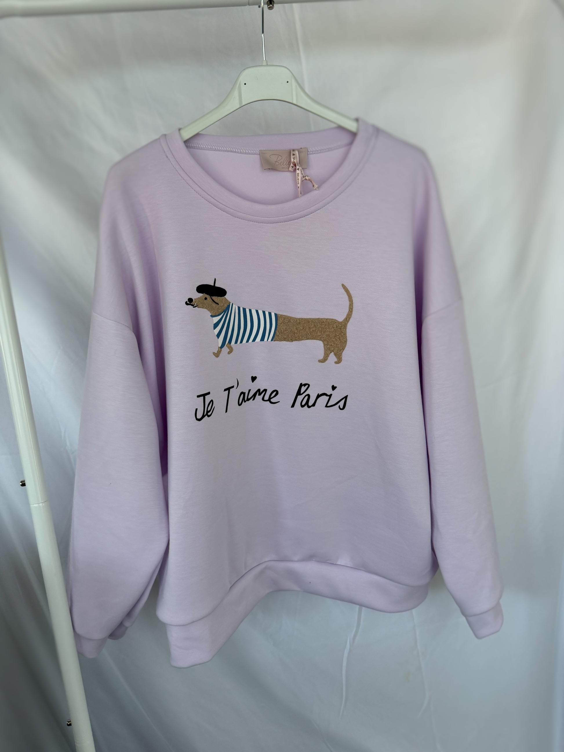 An einem weißen Kleiderbügel hängt ein hellvioletter Feinraummode Sweatshirt Dackelprint / Je t'aime Paris Schriftzug Flieder, der einen niedlichen Dackel mit Baskenmütze und gestreiftem Hemd zeigt, darunter "Je T'aime Paris".
