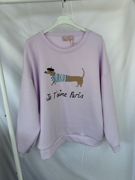 An einem weißen Kleiderbügel hängt ein hellvioletter Feinraummode Sweatshirt Dackelprint / Je t'aime Paris Schriftzug Flieder, der einen niedlichen Dackel mit Baskenmütze und gestreiftem Hemd zeigt, darunter "Je T'aime Paris".