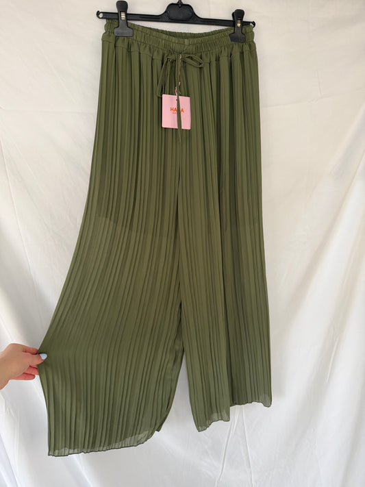 Die Plissee Hose Amanda von Feinraummode in Olivgrün mit elastischem Taillenbund und weitem Bein hängt an einem schwarzen Bügel vor weißem Hintergrund. Ein pinkes Etikett am Bund und eine Hand zeigen den fließenden Stoff.