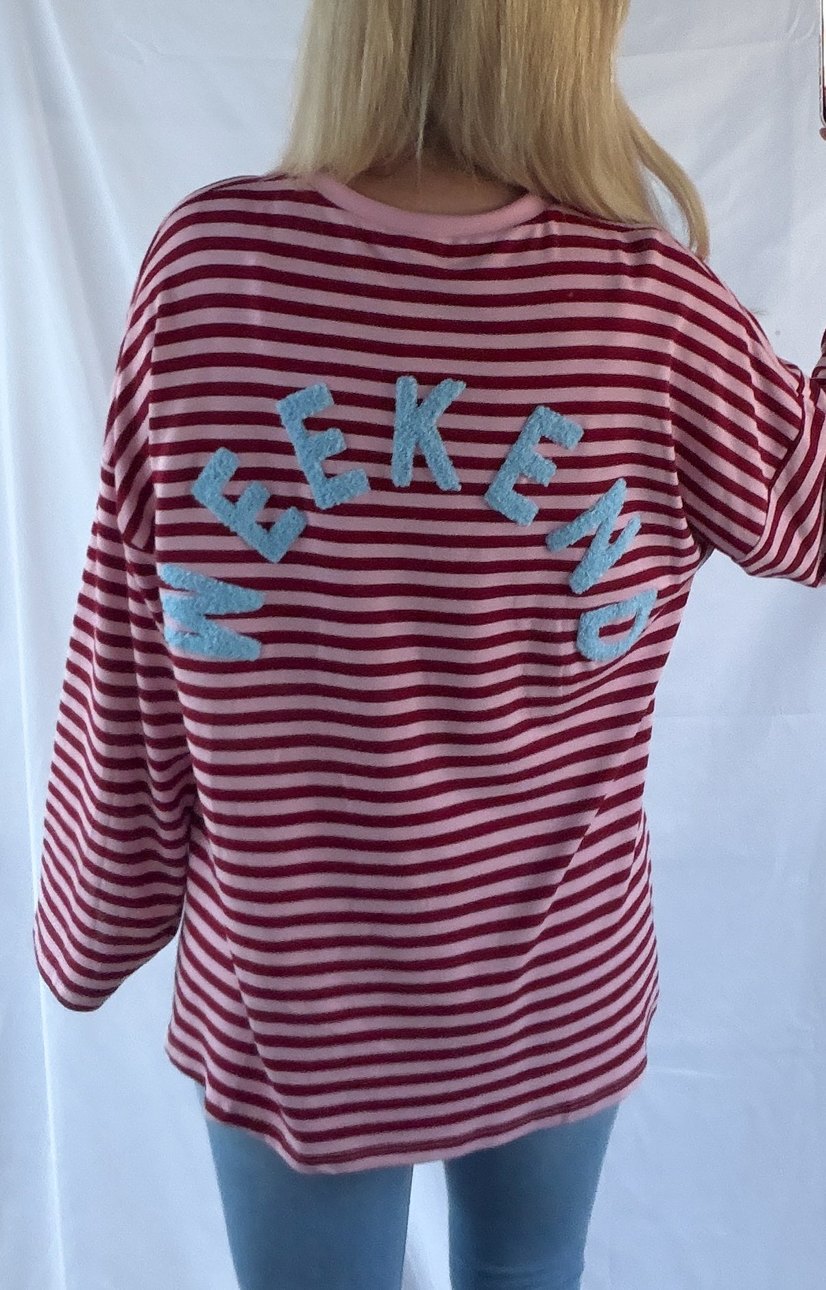 Von hinten sieht man eine Person mit langen blonden Haaren, die das Feinraummode Oversize Shirt Damen Pink gestreift mit auffälligem WEEKEND-Print trägt; die rosa-weißen Streifen stechen als Teil eines trendigen Streetwear-Looks hervor.