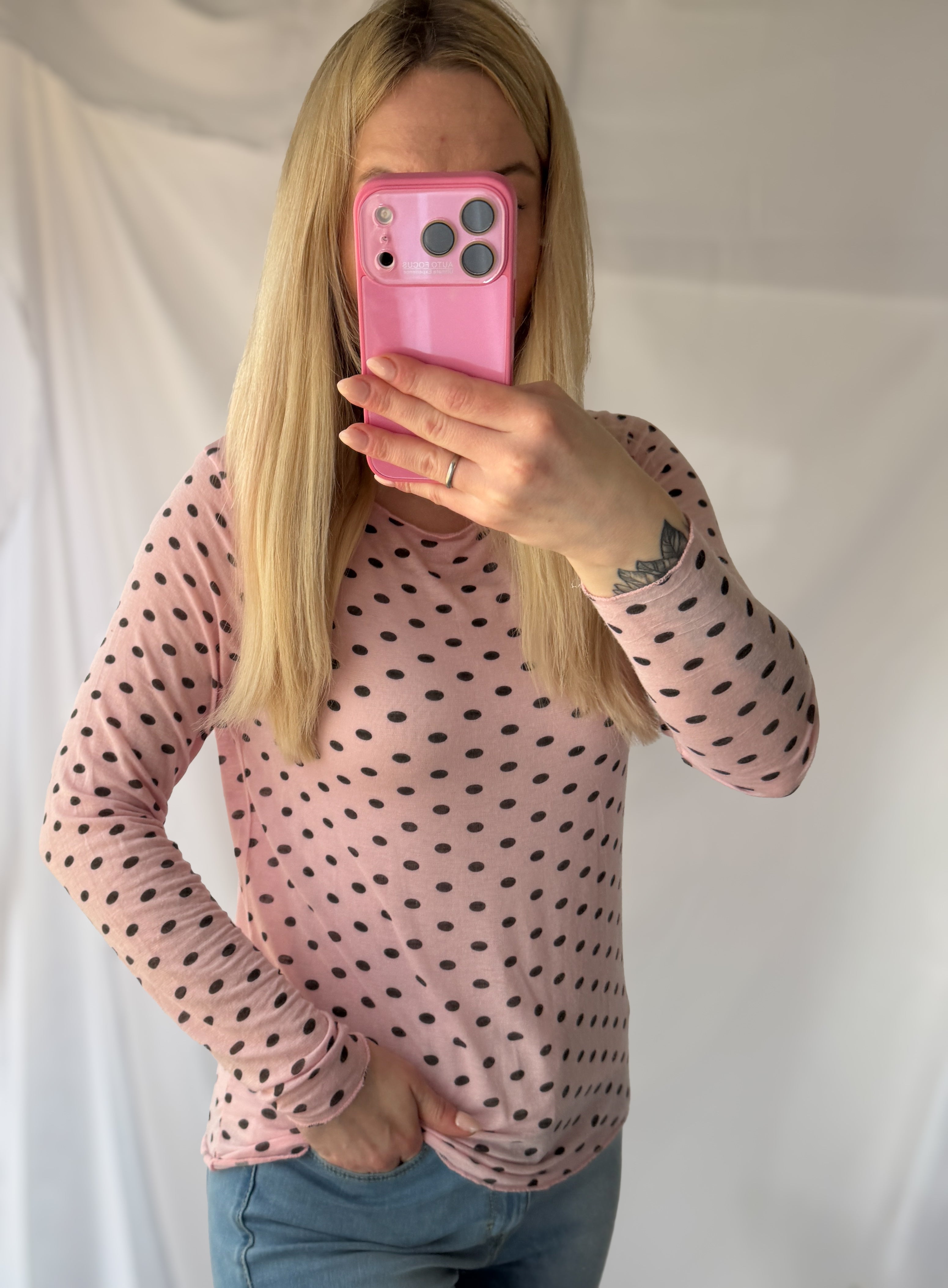 Eine Person mit langen blonden Haaren in der Feinraummode Gepunktetes Cashmere Longsleeve - Polka Dots rosa & wollweiß macht ein Spiegel-Selfie mit einem rosafarbenen Handy, ihr Gesicht teilweise verdeckt - ein schicker Moment in der zeitgenössischen Damenmode.
