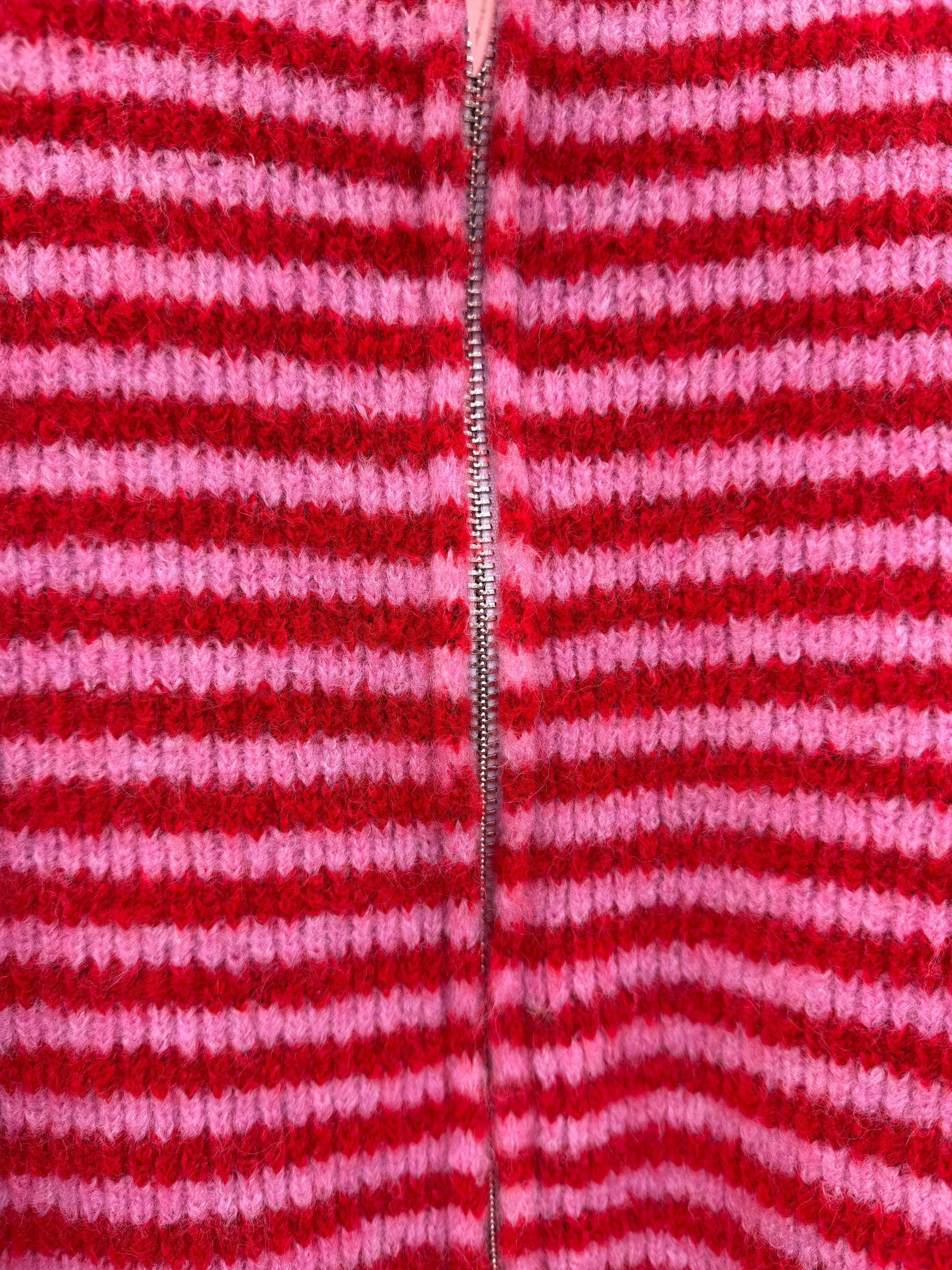 Nahaufnahme der Feinraummode Gestreifter Zip Cardigan rot: Gestrickter Stoff mit horizontalen roten und rosa Streifen und einem Metallreißverschluss in der Mitte.