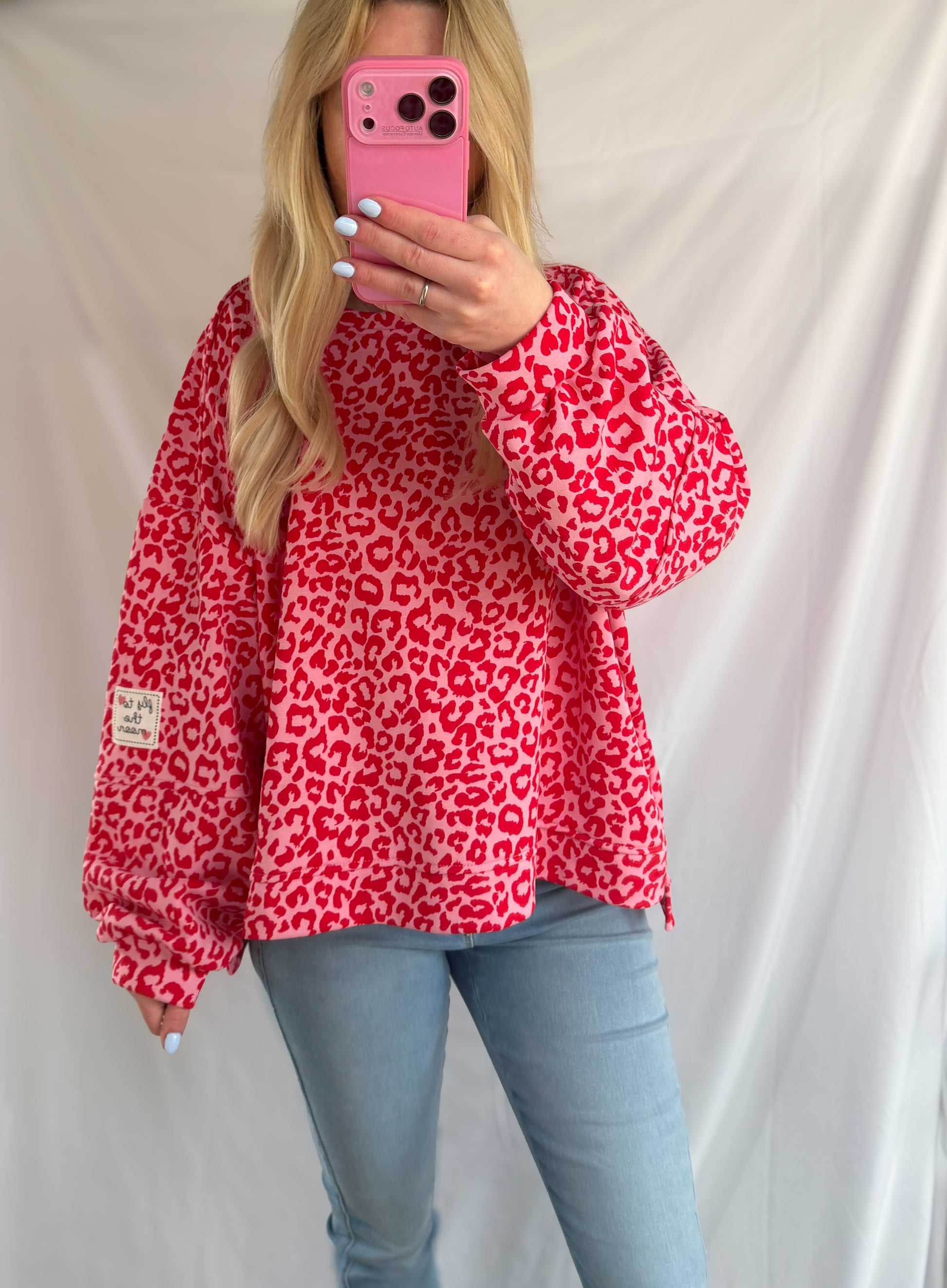 Eine Frau mit langen blonden Haaren macht ein Selfie im Spiegel. Sie trägt das Feinraummode Oversize Sweatshirt im Leoparden Look - rot, hellblaue Jeans und hält ein pinkes Handy in der Hand. Ihre Nägel sind weiß und der Hintergrund ist ein schlichtes weißes Laken.