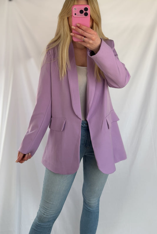 Eine Frau mit langen blonden Haaren trägt den Feinraummode Eleganter Nice Fit Blazer in flieder mit Leopardenprint, gepaart mit einem weißen Top und hellblauen Jeans. Sie steht vor einem weißen Hintergrund und macht ein Spiegel-Selfie mit einem pinken Handy.