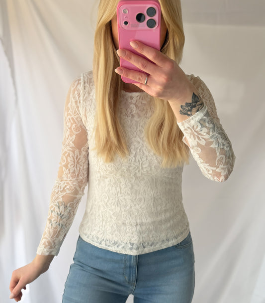 Eine Person mit langen blonden Haaren trägt die Feinraummode Shirt mit Spitze Pia in braun und weiß und hellblaue Jeans und macht ein Spiegel-Selfie mit einem pinkfarbenen Handy. Ihr Gesicht ist verdeckt und auf ihrem linken Handgelenk befindet sich ein Blumentattoo - elegante Damenmode pur.
