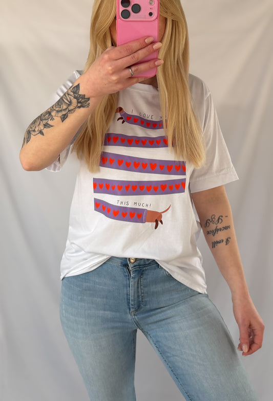 Eine Frau mit langen blonden Haaren trägt ein Feinraummode Weißes T-Shirt mit Dackelprint & Herzen, blaue Jeans und hält ein rosafarbenes Telefon, das ihr Gesicht teilweise verdeckt. Sie hat ein Blumentattoo auf ihrem linken Arm.