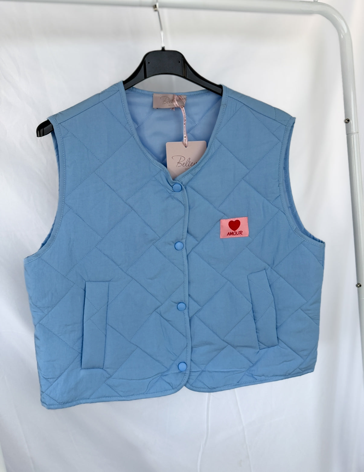 Die hellblaue Steppweste Ilona von Feinraummode präsentiert sich mit Druckknöpfen, pinkem Patch mit rotem Herz und "AMOUR", sowie "Belle"-Etikett am Kragen - ideal als Übergangsjacke Damen im Frühling.