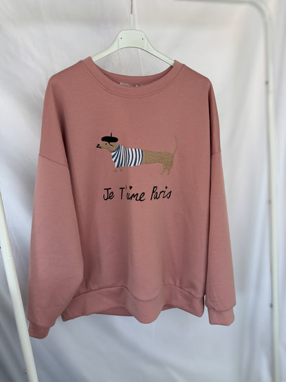 An einem weißen Kleiderbügel hängt ein rosafarbenes "Sweatshirt Dackelprint / Je t'aime Paris Schriftzug rosa" von Feinraummode, das einen gestreiften Dackel mit Baskenmütze und den Text "Je t'aime Paris" vor einem schlichten weißen Stoffhintergrund zeigt.