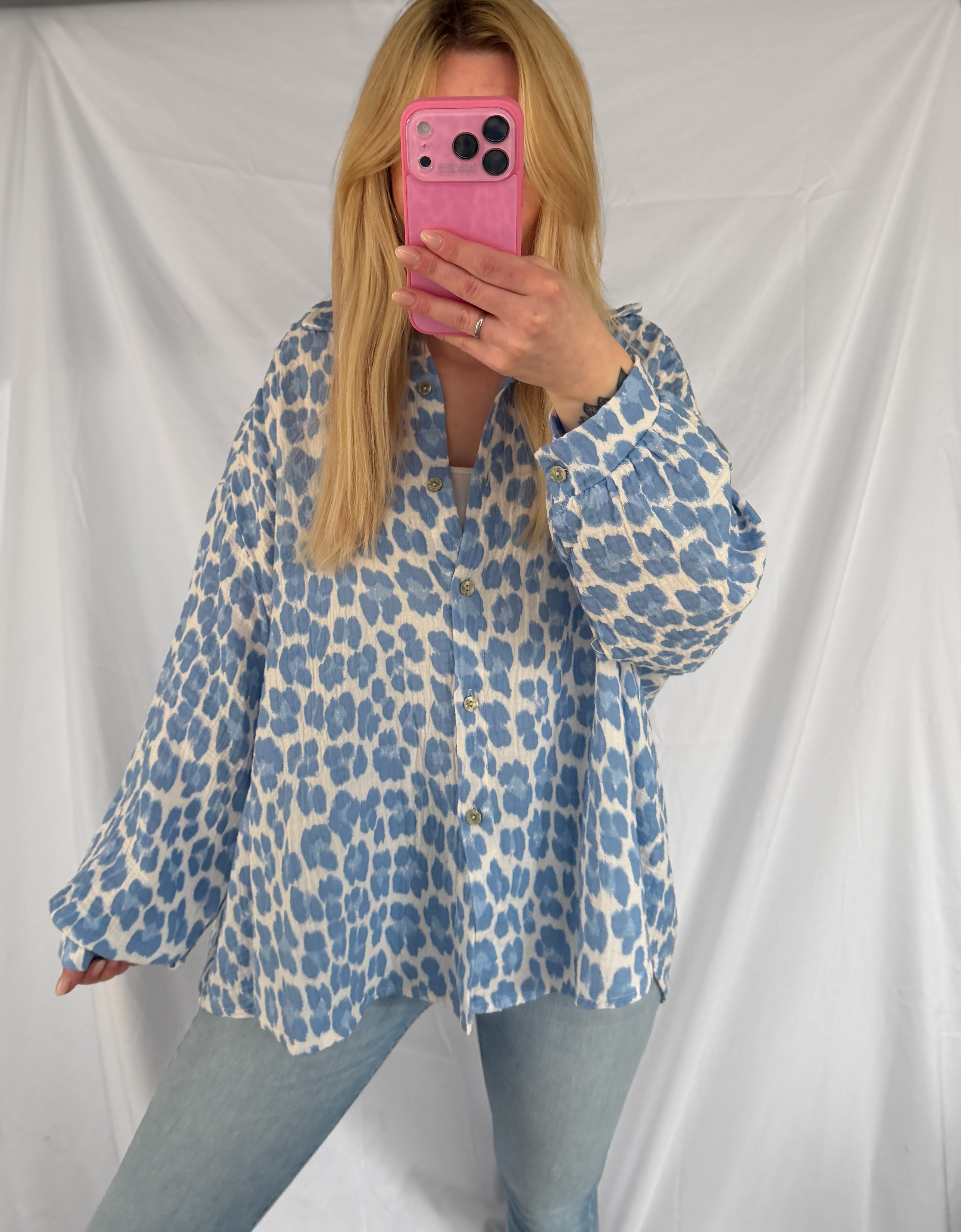 Eine Person mit langen blonden Haaren macht ein Selfie im Spiegel. Sie trägt die Oversize Musselinbluse Leopardenmuster blau weiß von Feinraummode und hellblaue Jeans und hält ein rosa Telefon in der Hand. Im Hintergrund ist ein weißes Laken zu sehen.