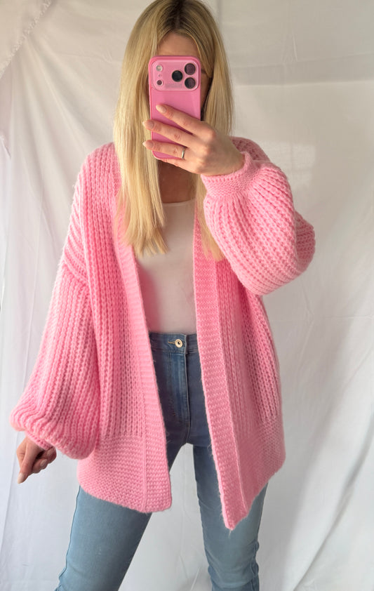 Eine Person mit langen blonden Haaren trägt den Kuscheliger Cardigan Lara in Pink von Feinraummode, ein weißes Oberteil und eine blaue Jeans und macht ein Spiegel-Selfie mit einem pinken Handy vor einem weißen Stoffhintergrund - ideal für Frühling & Herbst.
