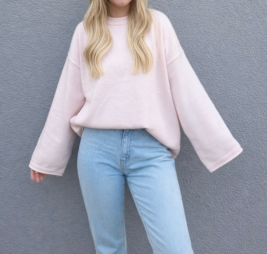 Eine Person steht vor einer grauen, strukturierten Wand und trägt den Cozy Oversize Sweater Mandy - Oversize Pullover Damen von Feinraummode in Hellrosa mit hochgezogenen hellblauen Jeans. Ihr lockeres, gewelltes blondes Haar ergänzt den gemütlichen Damenmode-Stil. Das Gesicht ist nicht sichtbar.
