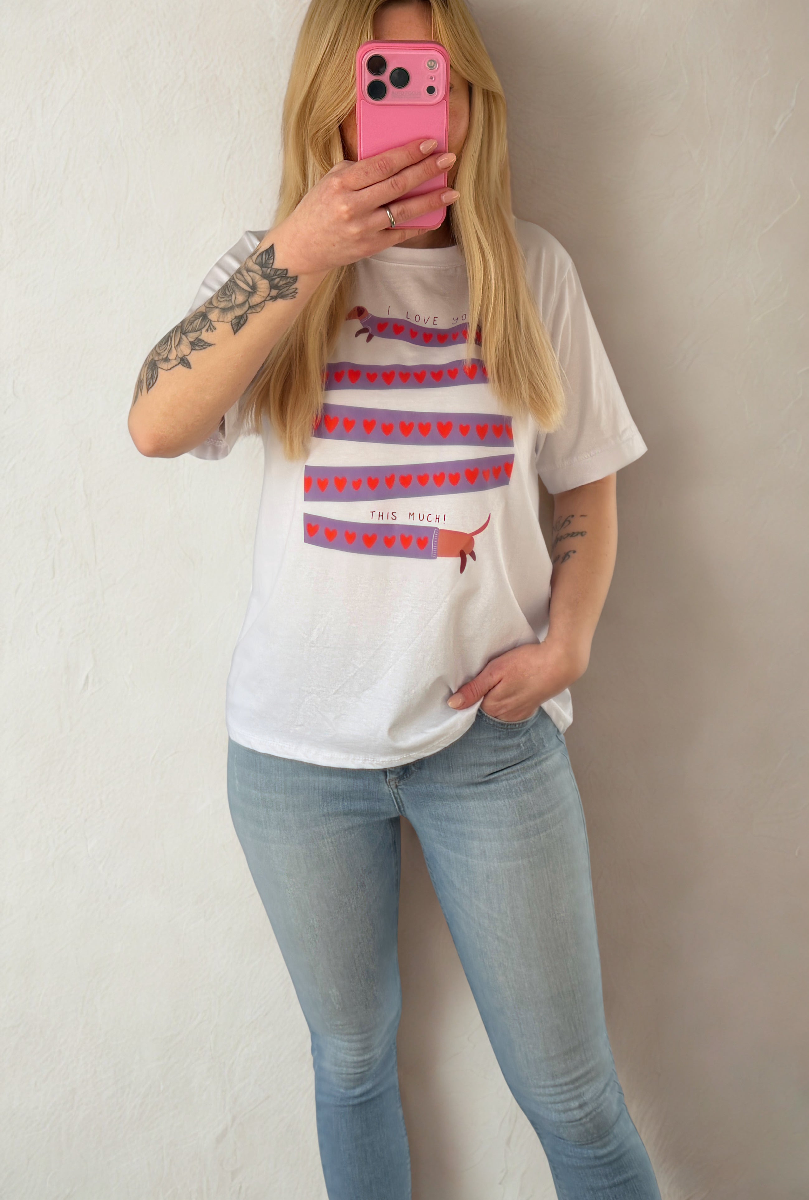 Eine Person mit langen blonden Haaren trägt die Feinraummode Weißes T-Shirt mit Dackelprint & Herzen und hellblaue Jeans, hält sich ein rosa Telefon vor das Gesicht und steht vor einer hellen Wand.